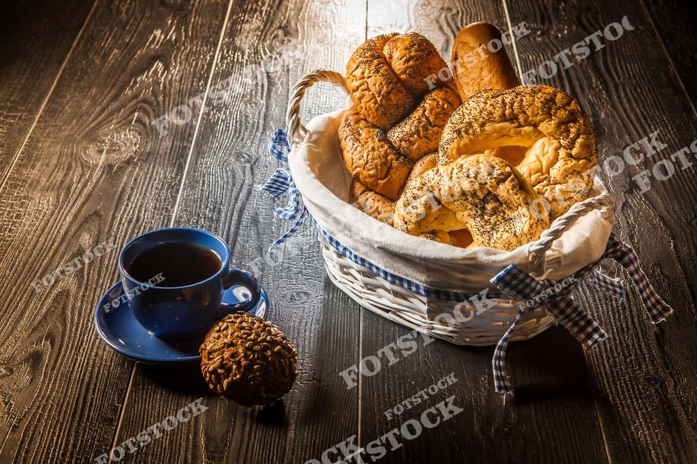 Натюрморт из продуктов питания. Рядом с чашкой кофе лежит цельнозерновая булочка. Также на столе размещена корзинка со свежей выпечкой.